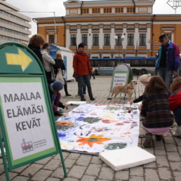 Ihmisiä Turun kauppatorilla tekemässä maalausta sekä kyltti 