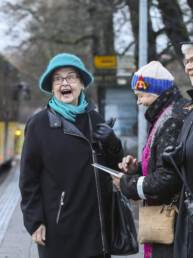 Kuvassa nauravia, iäkkäitä rouvia bussipysäkillä, taustalla linja-auto.