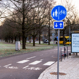 Pyörä- ja kävelytie, etualalla tietä kuvaava liikennemerkki, tien vieressä bussipysäkki ja taka-alalla linja-auto