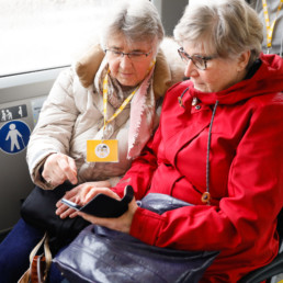 Kaksi harmaahiuksista naista istuu bussin penkillä ja katsovat kännykkää yhdessä. He ovat fölikavereita.