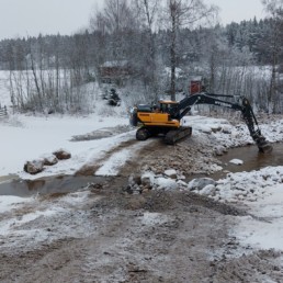Kuvassa kaivinkone rakenta uutta pohjapatoa lumisessa maisemassa.