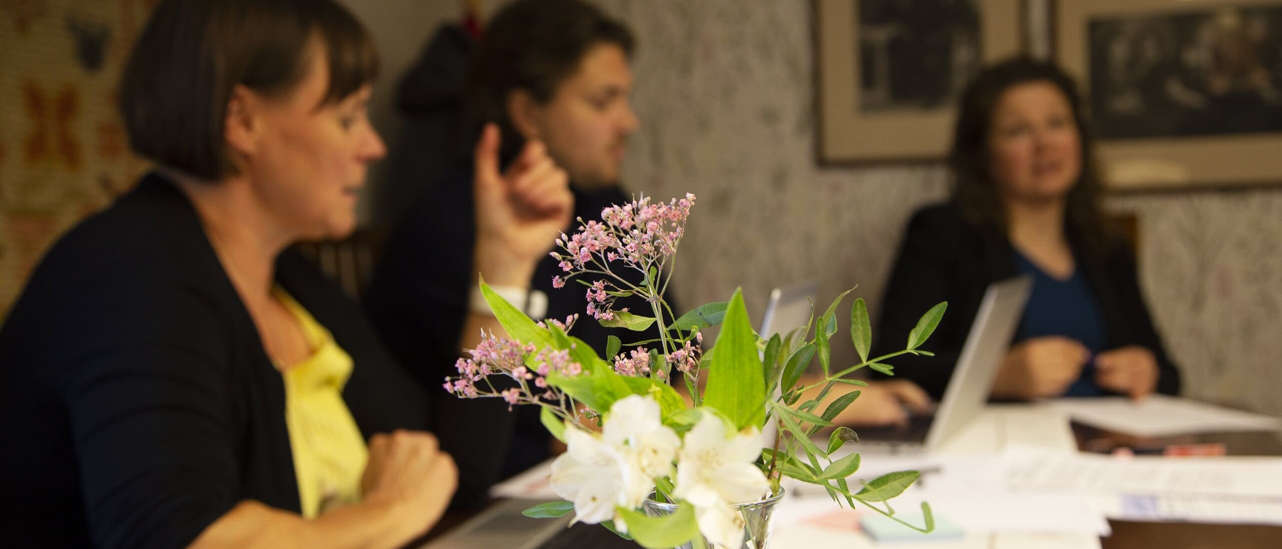 Kolme ihmistä keskustelemassa pöydän ympärillä, etualalla kukkamaljakko.