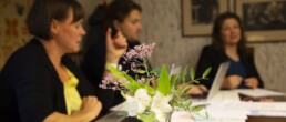 Kolme ihmistä keskustelemassa pöydän ympärillä, etualalla kukkamaljakko.
