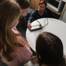 Kolme oppilasta ja opettaja pöydän ympärillä. Yksi oppilaista puhuu mikrofoniin.