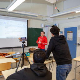 Kuva luokkahuoneesta, jossa päättäjä luokan edessä puhumassa oppilaille ja kaksi oppilasta kuvaamassa tapahtumaa videolle.