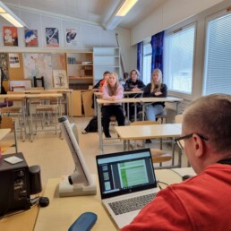 Kuntapäättäjä istuu koneen edessä luokkahuoneessa ja taustalla näkyy oppilaita pöytien ääressä kuuntelemassa.