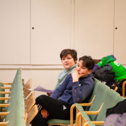 Kaksi oppilasta istuvat auditoriossa, toisella lappu kädessä, mietteiliään näköisiä