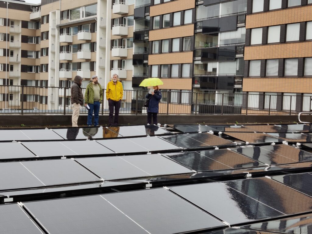Aurinkopaneeleja katolla, neljä ihmistä seisoo paneelien takana, taustalla kerrostalojen seiniä.