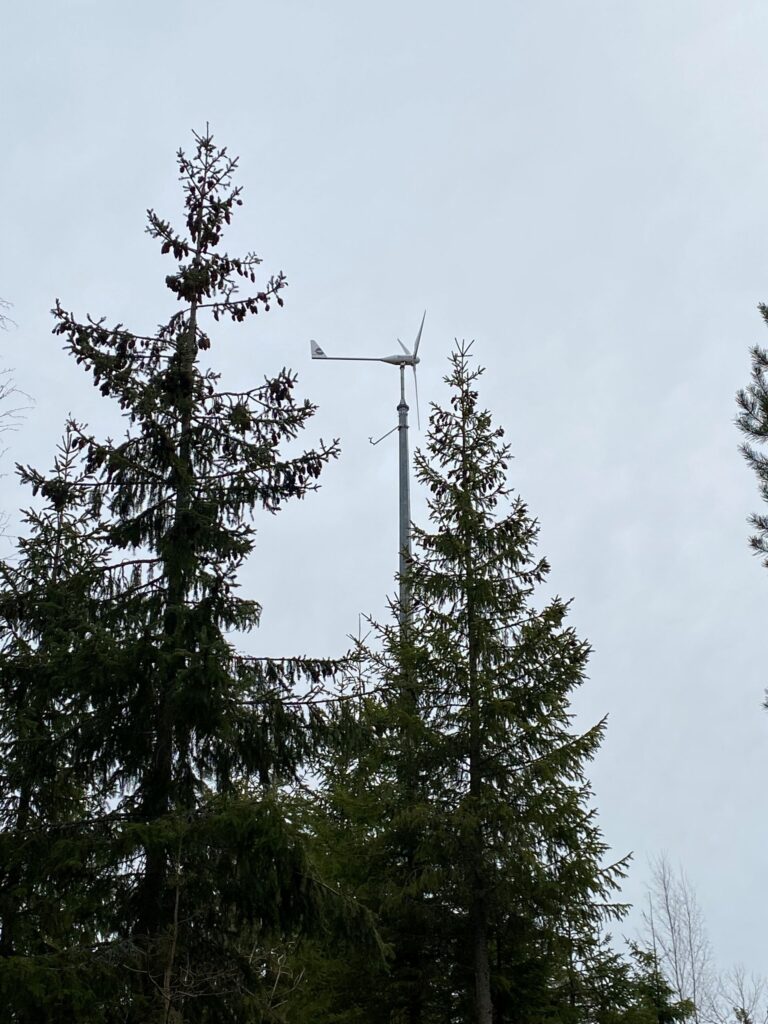 Pientuulivoimalan yläosa näkyy kahden kuusen latvan takana.
