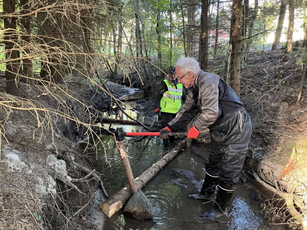 Henkilö ojassa puusuisteen edessä lyömässä lekaa vaarnaan.