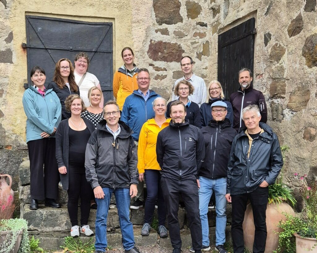 Joukko hymyileviä ihmisiä vanhan, muuratun kiviseinän edustalla Qvidjassa.