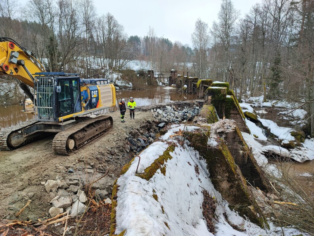 Kaivinkone ja kaksi henkilöä patorakennelman päällä kevättalvisena päivänä