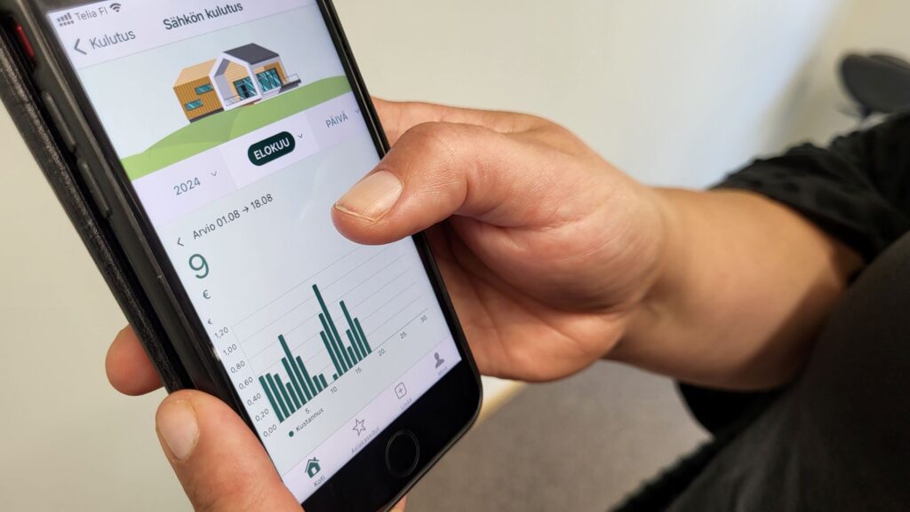 kädet pitelevät älypuhelinta jonka näytöllä sähkön kulutusseurantaa