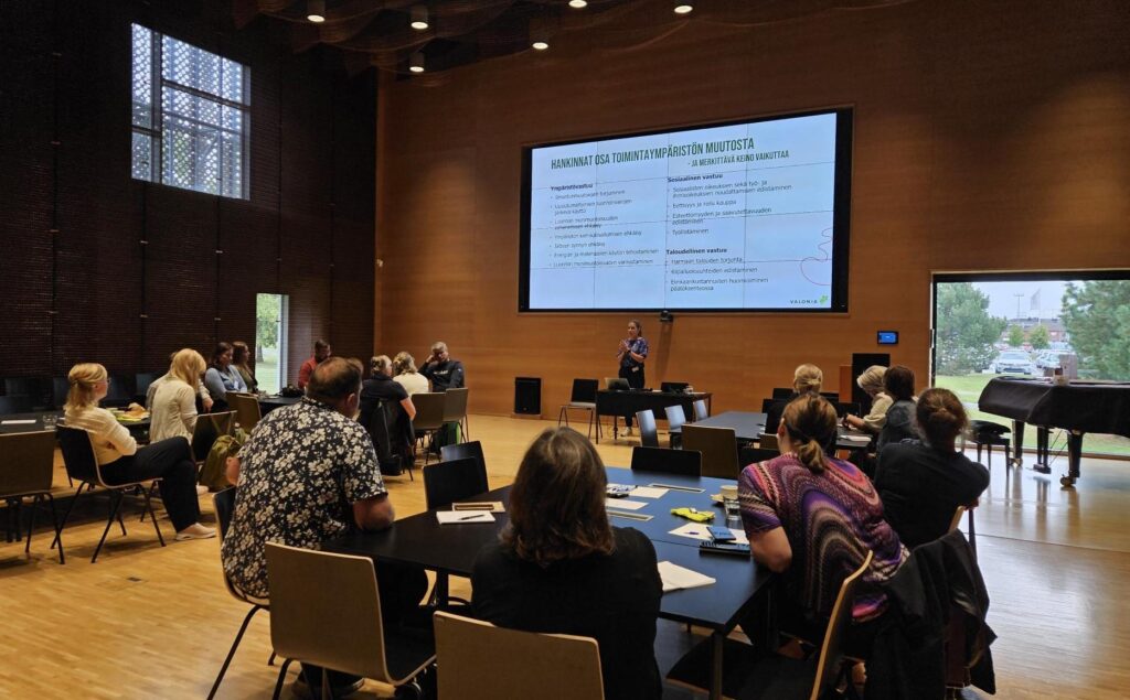 Sali, jossa kuntien edustajia pöytien ääressä ja puhuja edessä.