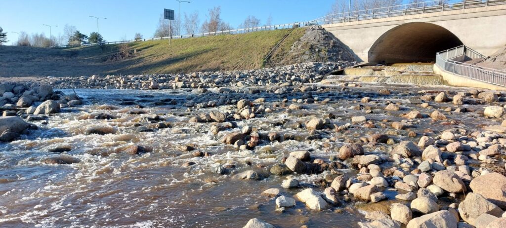 Koskimaisemaa, jossa paljon kiviä, taustalla betoninen silta ja ruohoista pengertä.
