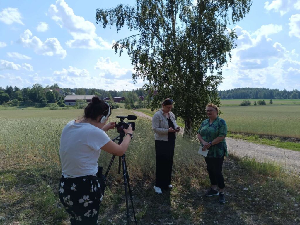 Kaksi henkilöä pellon laidalla, kolmas henkilö kuvaa heitä videokameralla.
