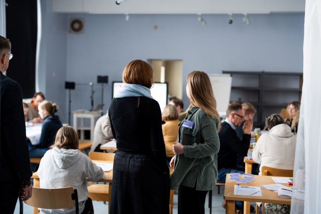 ihmisiä työpajassa sisällä, kaksi seisoo etualalla, muut istuvat pöydissä