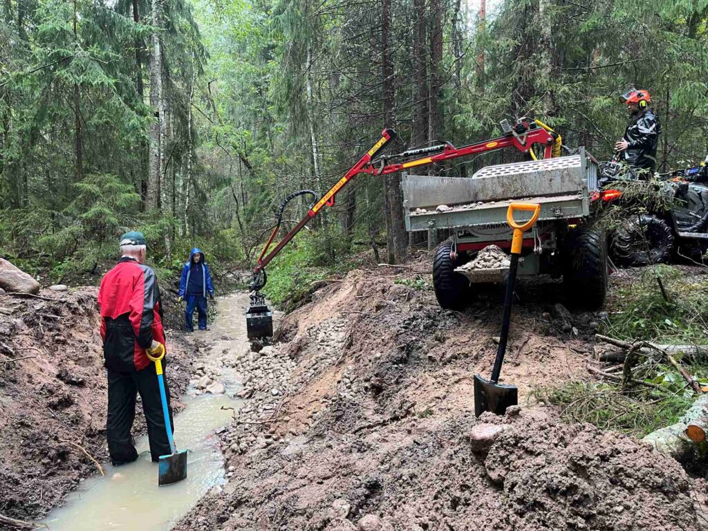 Kaksi henkilöä purossa, puron reunalla mönkijän peräkärry ja kuormaaja joka nostelee isoja kiviä puron pohjalle.