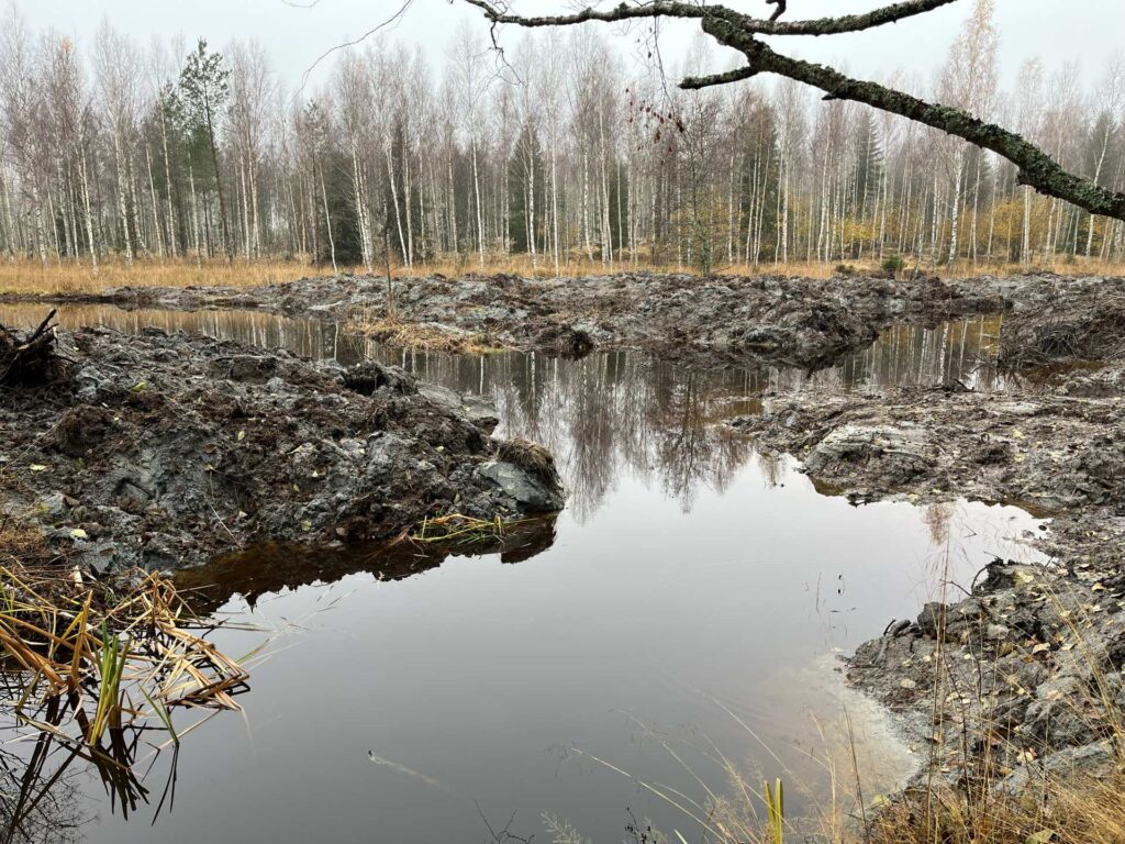 Keskellä uoma, jossa vettä. Uoman sivuilla saviset penkereet. Taustalla puustoa.