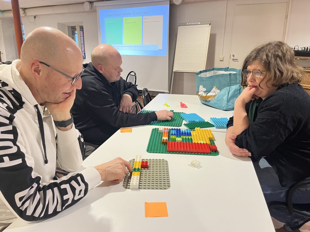 Kolme henkilöä istuu pöydän äärellä ja katselee viljelysuunnitelmaa legopalikoista