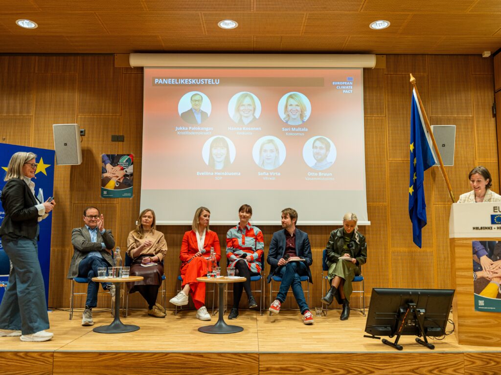 Lavalla istuu paneelikeskustelu: kuusi osallistujaa tuoleilla pöytien ääressä. Taustalla valkokangas, jossa lukee ‘Paneelikeskustelu’ ja on puhujien kuvat ja nimet. Sivulla EU-liput ja edessä puhujapönttö.