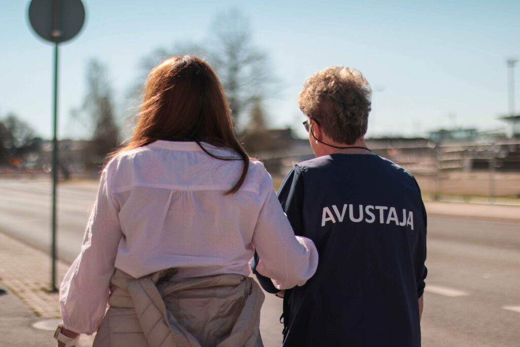 Kuvassa takaantapäin kuvattuna vasemmalla näkövammainen ja oikealla hänen vieressään avustaja. He kulkevat kadulla.
