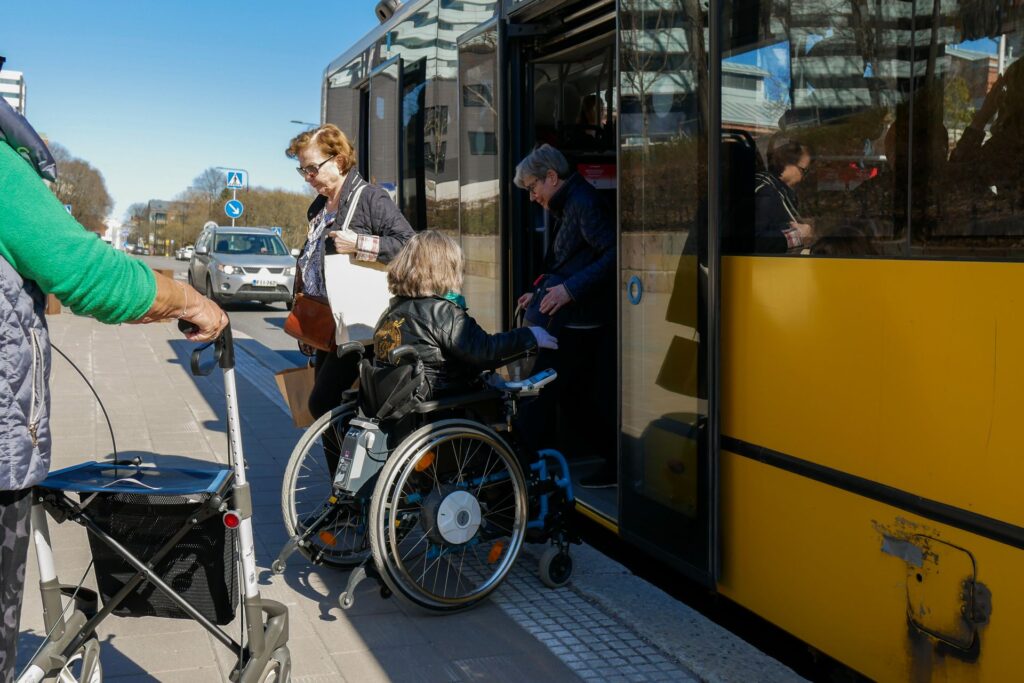 Pyörätuolia käyttävä osallistuja nousee bussiin muiden osallistujien vieressä esteettömyyden kokeilutilanteessa.
