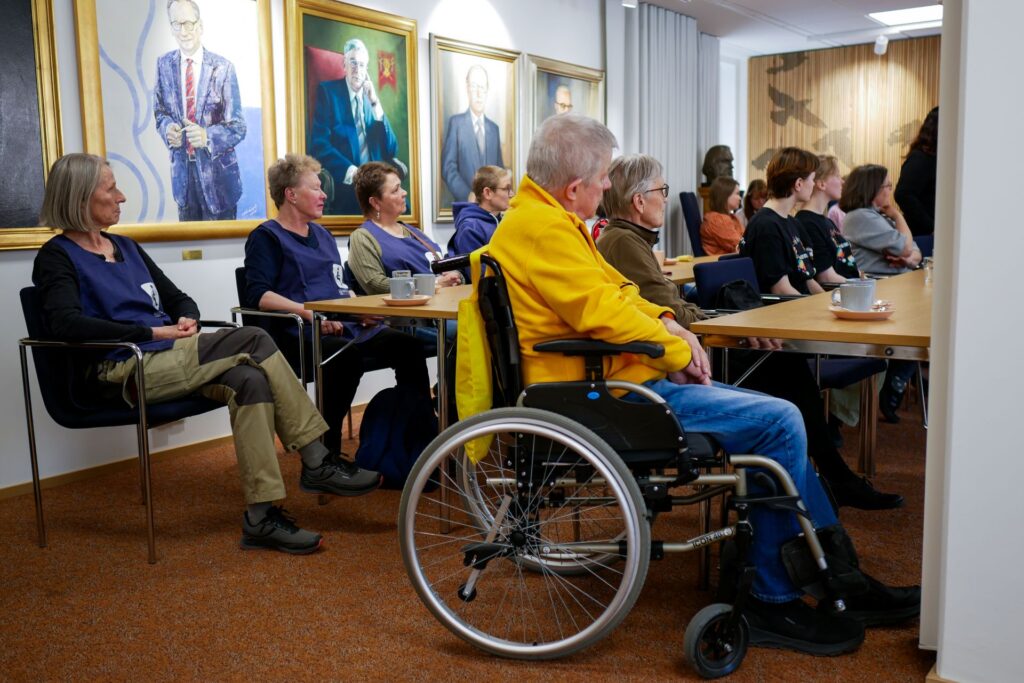 Osallistujat seuraavat tilaisuutta istuen pöytien ääressä, etualalla pyörätuolia käyttävä henkilö.