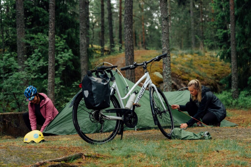 Etulalla pyörä jossa tarakkalaukut, taka-alalla kaksi ihmistä pystyttää telttaa