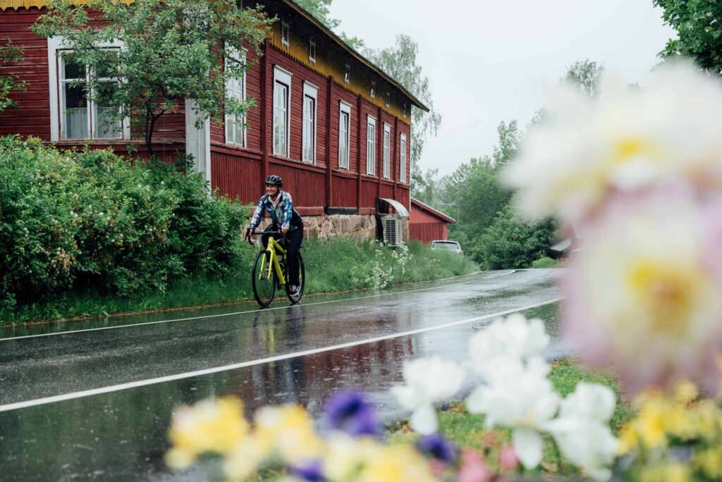 Pyöräilijä sateisena kesäpäivänä maantiellä. Taustalla vanha, punainen puurakennus, etualalla kukkia