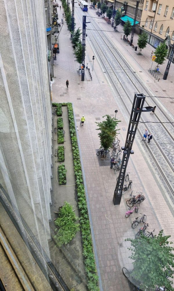 Näkymä ylhäältä vilkkaalle kadulle, jossa kulkee ratikkakiskot. Rakennuksen seinustalla on pitkänä nauhana istutusalueita, ja kadulla näkyy pyöräilijöitä ja jalankulkijoita.