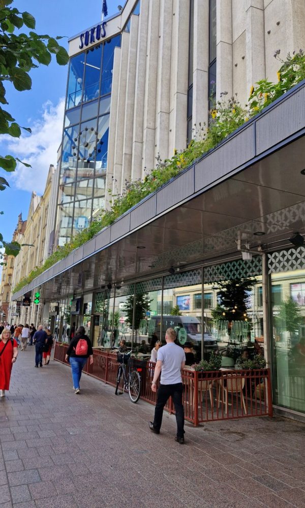 Kaupunkikatu, jonka varrella on korkea liikerakennus. Rakennuksen katosrakenteen päällä kasvaa pitkä vihreä istutusalue. Kadulla kävelee ihmisiä ja muutama polkupyörä on pysäköity.