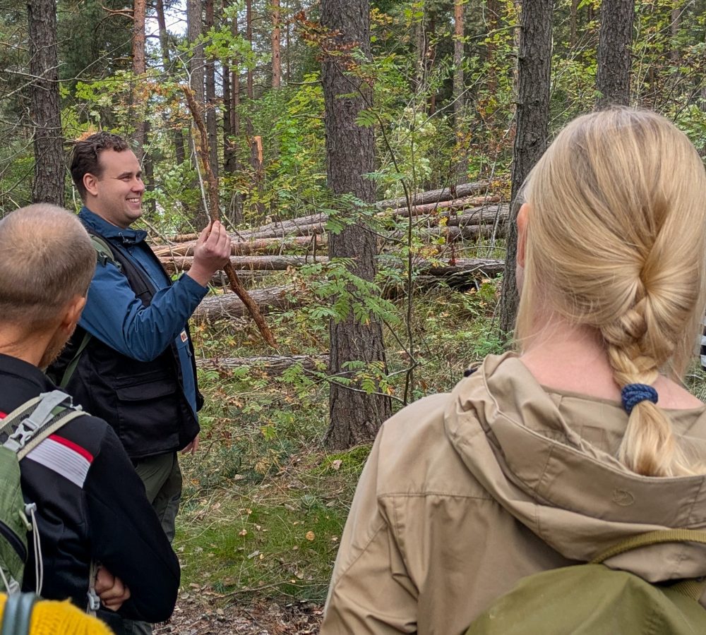 Ihmisiä metsässä. Hymyilevä ihminen keppi kädessä.