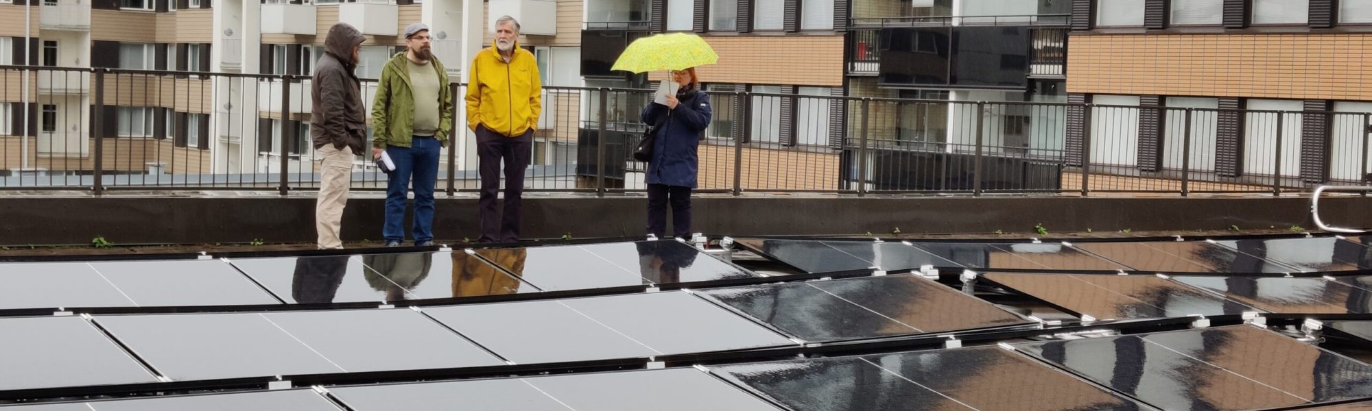 Aurinkopaneeleja katolla, neljä ihmistä seisoo paneelien takana, taustalla kerrostalojen seiniä.